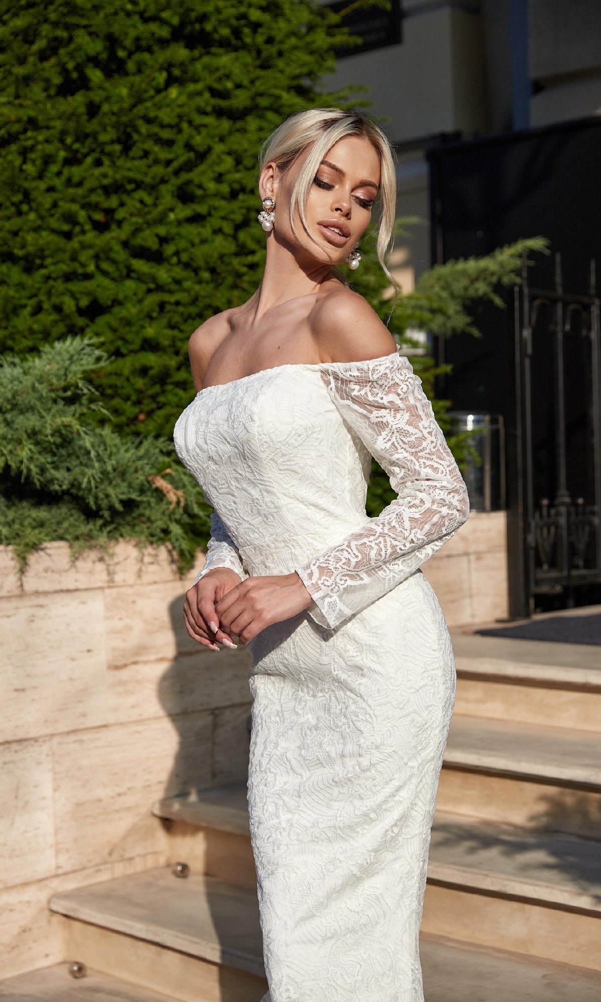 A woman in the Long Lace White Formal Gown: Tina Holly BB007, featuring an off-the-shoulder design and long lace sleeves, stands outdoors near stone steps and lush greenery.